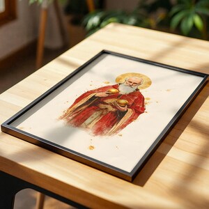 May include: A framed print featuring a watercolour-style illustration of a bearded figure with a golden halo, wearing a red robe and holding two golden spheres. The artwork is set against a cream-coloured background with subtle gold paint splatters.