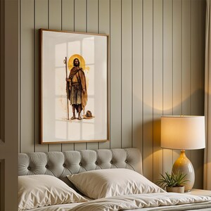 May include: A framed wall art print featuring a standing figure with a golden halo, wearing a brown robe and holding a tall wooden staff. The figure is depicted in a watercolour style with a small hat resting on the ground near their feet.