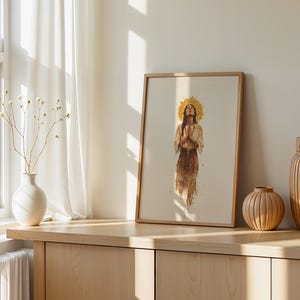 May include: A framed vertical wall art print featuring a watercolour illustration of a person with long dark hair, hands pressed together in prayer, and a golden sunburst halo behind their head, rendered in earthy brown and tan tones.