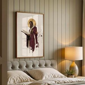 May include: A framed wall art print featuring a bearded figure in a deep maroon robe holding an open book, accented by a gold circular halo and dark ink-like splatters on a white background, displayed in a bedroom with a tufted headboard.