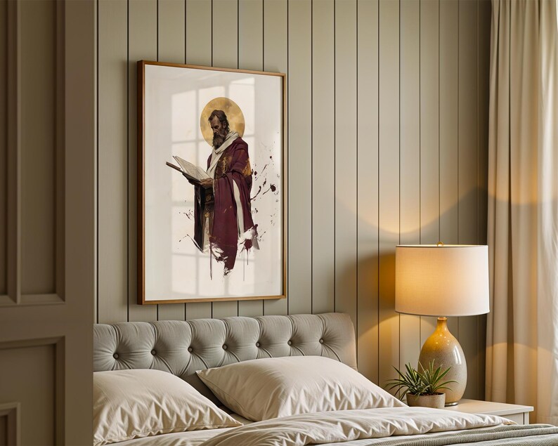 May include: A framed wall art print featuring a bearded figure in a deep maroon robe holding an open book, accented by a gold circular halo and dark ink-like splatters on a white background, displayed in a bedroom with a tufted headboard.