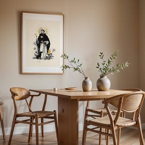 Op de afbeelding: Een ingelijste wandkunstprint met een zwart-witillustratie van een figuur in een gewaad met een gouden halo, een kleine hond en florale elementen. De print is ingelijst in een lichthouten lijst en hangt aan een beige muur boven een eettafel.