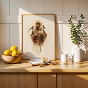 May include: A framed watercolour-style print depicting a bearded figure in a brown robe with a golden halo, holding white flowers with birds perched on the shoulders, displayed alongside a bowl of yellow lemons and a white vase with green leaves.