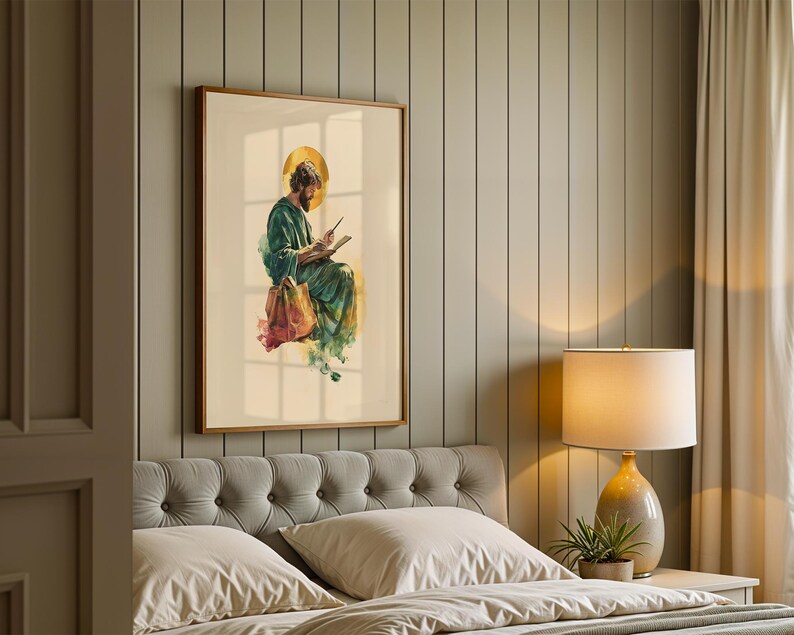 May include: A framed wall art print featuring a watercolor illustration of a bearded figure in a green robe with a gold halo, writing in a book. The figure sits with a brown bag at their feet, rendered in soft, flowing brushstrokes on a cream background.