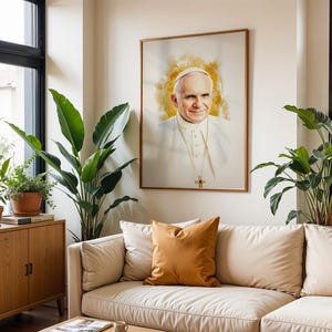 May include: A framed portrait of Pope John Paul II featuring a watercolour-style golden halo effect behind the head. The subject wears a white cassock and a gold pectoral cross. The artwork is displayed on a wall above a beige sofa with pillows.