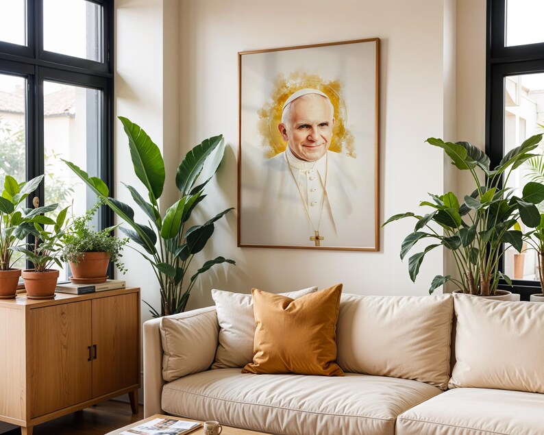 May include: A framed portrait of Pope John Paul II featuring a watercolour-style golden halo effect behind the head. The subject wears a white cassock and a gold pectoral cross. The artwork is displayed on a wall above a beige sofa with pillows.