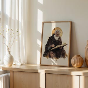 Peut inclure: Une impression d'art encadr&eacute;e repr&eacute;sentant une personne &acirc;g&eacute;e &agrave; barbe blanche v&ecirc;tue d'une robe &agrave; capuche marron, assise et &eacute;crivant dans un livre ouvert avec une aur&eacute;ole dor&eacute;e derri&egrave;re la t&ecirc;te, pr&eacute;sent&eacute;e dans un cadre en bois clair.