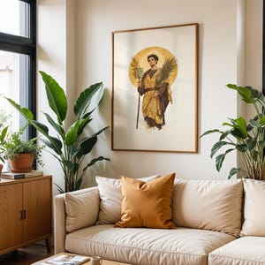 Op de afbeelding: Een ingelijste kunstdruk met een klassieke illustratie van een figuur in gouden gewaden die een palmtak en een staf vasthoudt, tegen een achtergrond van een cirkelvormige gouden halo, weergegeven in een kamer met een beige bank en groene planten.
