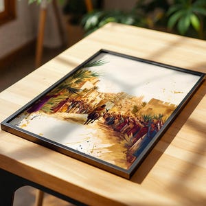 May include: A framed rectangular art print featuring a watercolour-style illustration of a crowd of people in robes lining a dusty path. A figure rides a dark donkey towards a stone city gate under palm trees, rendered in warm earth tones and gold.