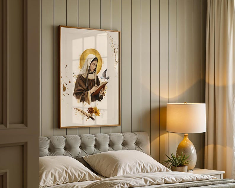 May include: A framed wall art print featuring a figure in a brown habit and white head covering, holding a book and pen with a golden halo behind the head. A small bird flies nearby, and gold paint splatters decorate the white background.