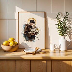 Puede incluir: Una impresi&oacute;n enmarcada de estilo acuarela que representa una figura con h&aacute;bito marr&oacute;n sosteniendo a un ni&ntilde;o peque&ntilde;o, con un halo dorado, un lirio blanco y un libro. La obra presenta salpicaduras de pintura azul y marr&oacute;n sobre fondo blanco.