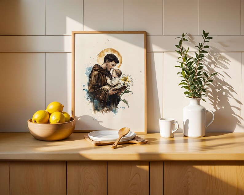 Puede incluir: Una impresi&oacute;n enmarcada de estilo acuarela que representa una figura con h&aacute;bito marr&oacute;n sosteniendo a un ni&ntilde;o peque&ntilde;o, con un halo dorado, un lirio blanco y un libro. La obra presenta salpicaduras de pintura azul y marr&oacute;n sobre fondo blanco.