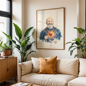 May include: A framed wall art print featuring a watercolor-style portrait of a bearded man with a halo, wearing blue vestments and holding a glowing, fiery red heart at the center of his chest, displayed in a light-colored interior space.