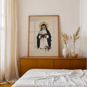 May include: A framed wall art print featuring a black and white illustration of a figure wearing a dark habit and a crown of thorns, holding an open book with red pages, set against a gold halo, with ink-like splatters at the bottom edge.
