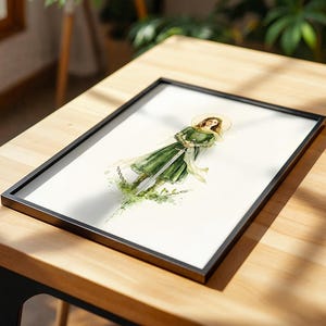 Op de afbeelding: Een ingelijste print met een aquarelillustratie van een figuur in een groene jurk die een zwaard vasthoudt. De figuur heeft een gouden halo achter het hoofd en is omringd door groen gebladerte en gebroken kettingen aan de onderzijde.