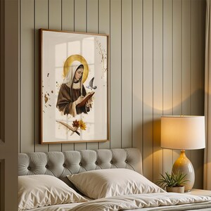 May include: A framed wall art print featuring a figure in a brown habit and white head covering, writing in a book with a gold halo behind their head. A small bird flies nearby, with gold paint splatters and floral accents below the figure.