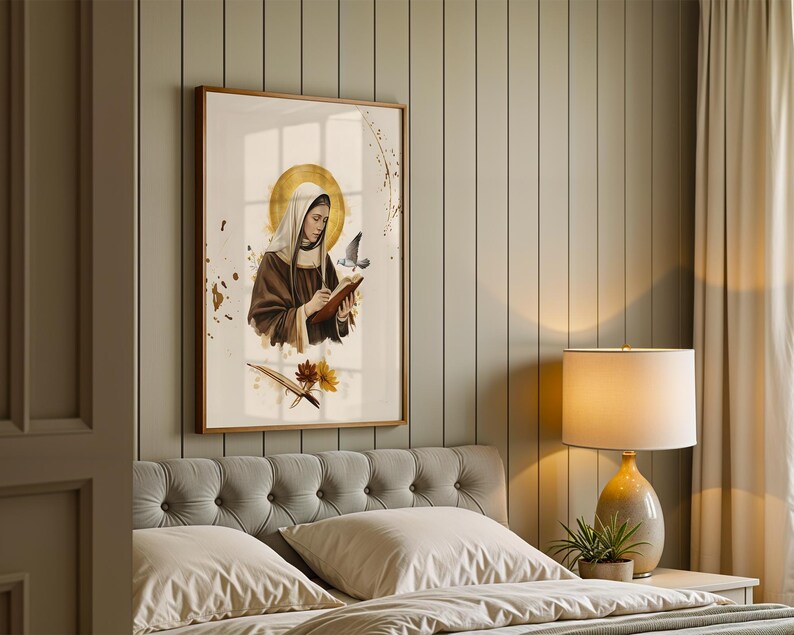 May include: A framed wall art print featuring a figure in a brown habit and white head covering, writing in a book with a gold halo behind their head. A small bird flies nearby, with gold paint splatters and floral accents below the figure.