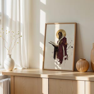 May include: A framed wall art print featuring a bearded figure in a deep maroon robe holding an open book, accented by a gold circular halo. The artwork displays a watercolour style with dark ink splatters on a light background, presented in a simple wooden frame.