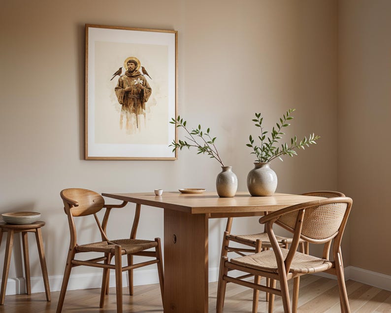 May include: A framed wall art print depicting a figure in brown robes with a golden halo, flanked by two birds, hanging above a wooden dining table with two light-colored ceramic vases containing green leafy branches and two wooden chairs.