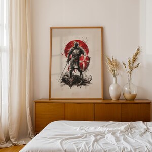May include: A framed wall art print featuring a knight in full armour holding a sword and a shield with a red cross, standing over a dark creature. The artwork is set against a large red circular background with black ink splatter accents.