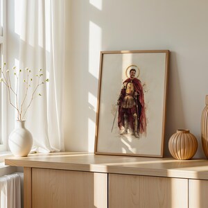 Puede incluir: Una l&aacute;mina de arte mural vertical enmarcada con una ilustraci&oacute;n en acuarela de una figura hist&oacute;rica con armadura dorada y una capa roja fluida, sosteniendo una espada. La figura tiene un halo dorado sobre un fondo de color crema claro.