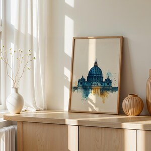 May include: A framed wall art print featuring a watercolour illustration of a large domed cathedral in shades of blue and gold. The artwork depicts the architectural structure with a reflective effect at the base, set against a light cream background.
