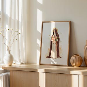 May include: A framed art print featuring a watercolor illustration of a figure in a brown habit and white robe, holding a bouquet of flowers and a small cross, with a golden halo, displayed in a light wood frame on a wooden surface.