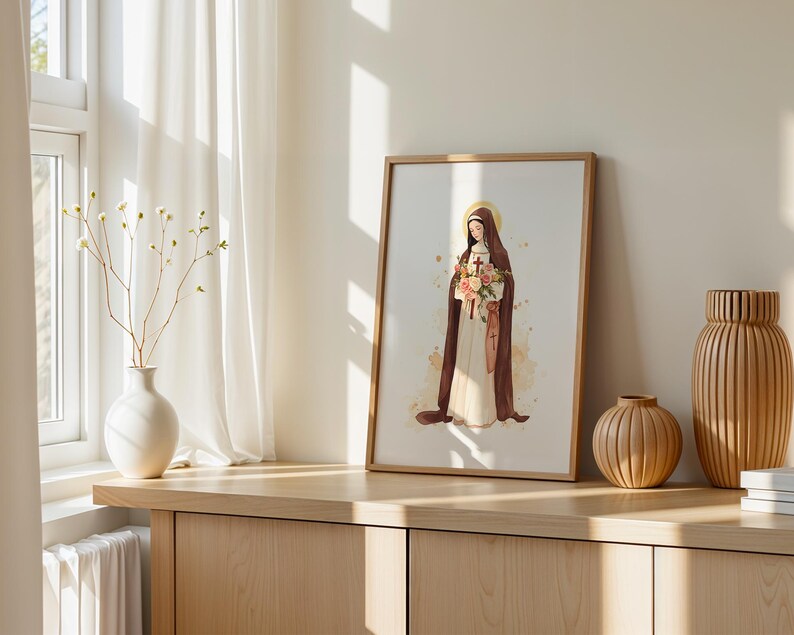 May include: A framed art print featuring a watercolor illustration of a figure in a brown habit and white robe, holding a bouquet of flowers and a small cross, with a golden halo, displayed in a light wood frame on a wooden surface.
