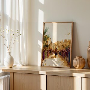 Peut inclure: Une impression encadr&eacute;e de style aquarelle repr&eacute;sentant une silhouette montant un &acirc;ne &agrave; travers une foule dans une ville d&eacute;sertique, avec des tons terreux chauds, des palmiers et une architecture en pierre, pr&eacute;sent&eacute;e dans un cadre en bois clair.