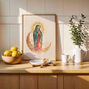 Puede incluir: Una l&aacute;mina de arte enmarcada con una ilustraci&oacute;n en acuarela de la Virgen de Guadalupe sobre una luna creciente con un halo dorado, expuesta junto a un cuenco de madera con limones amarillos, un plato blanco y un jarr&oacute;n blanco.