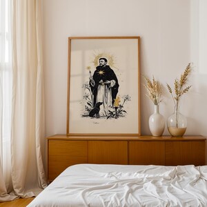 Op de afbeelding: Een ingelijste kunstdruk met een zwart-witillustratie van een figuur in een gewaad met een gouden sterrenhalo, die een kruis vasthoudt, vergezeld door een zwarte hond, lelies en een fakkel op een cr&egrave;mekleurige achtergrond.