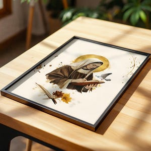 May include: A framed print featuring an illustration of a figure in a brown habit and white head covering writing in a book. A golden halo surrounds the head, with a small bird, a quill, and yellow and brown flowers depicted on a cream background.
