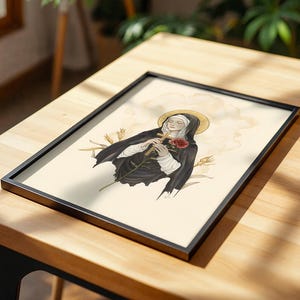 Op de afbeelding: Een ingelijste kunstdruk met een illustratie van een figuur in een zwart habijt en witte wimpel, die een houten kruis en een rode roos vasthoudt. De figuur heeft een gouden aureool en de achtergrond bevat korenaren en lichte wolken.