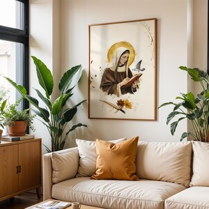 May include: A framed wall art print featuring a figure in a brown habit and white head covering writing in a book. A golden halo surrounds the head, with a small bird nearby. Below, a quill pen and two flowers are depicted on a cream background.