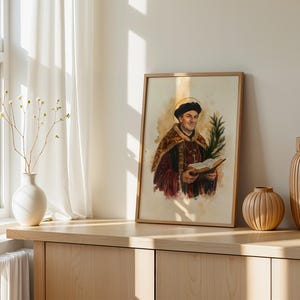May include: A framed watercolor print depicting a historical figure wearing a dark cap and a fur-trimmed robe, holding an open book and a palm frond. The figure has a golden halo behind their head, set against a light, neutral-toned background.