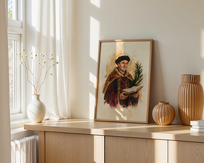 May include: A framed watercolor print depicting a historical figure wearing a dark cap and a fur-trimmed robe, holding an open book and a palm frond. The figure has a golden halo behind their head, set against a light, neutral-toned background.