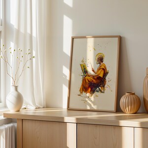 May include: A framed art print featuring a watercolour-style illustration of a bearded figure in orange robes sitting on a chair and painting on a small canvas. The artwork includes a golden halo and yellow paint splatters on a cream background.
