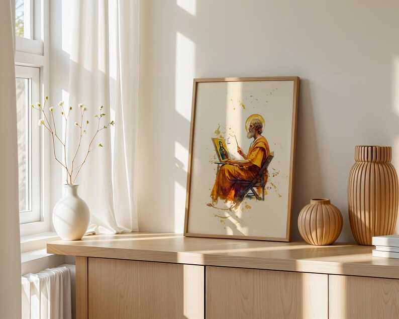 May include: A framed art print featuring a watercolour-style illustration of a bearded figure in orange robes sitting on a chair and painting on a small canvas. The artwork includes a golden halo and yellow paint splatters on a cream background.
