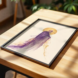 May include: A framed art print featuring an illustration of a figure in a flowing purple robe with a golden halo behind the head. The artwork is set against a cream-coloured background, presented in a thin black frame on a wooden surface.