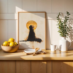 May include: A framed art print featuring a figure in a black hooded robe sitting beneath a large golden moon. The artwork is displayed on a counter next to a wooden bowl of yellow lemons, a stack of white plates, a wooden spoon, and a white vase.