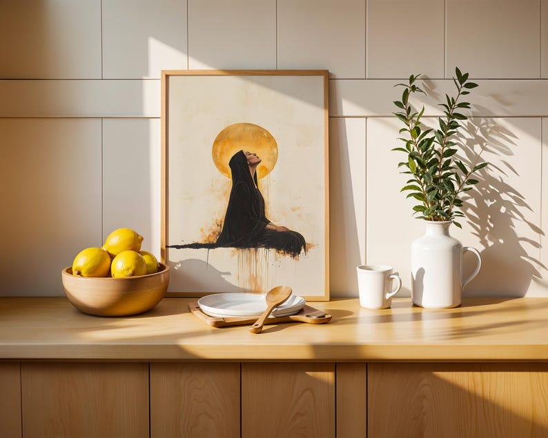 May include: A framed art print featuring a figure in a black hooded robe sitting beneath a large golden moon. The artwork is displayed on a counter next to a wooden bowl of yellow lemons, a stack of white plates, a wooden spoon, and a white vase.