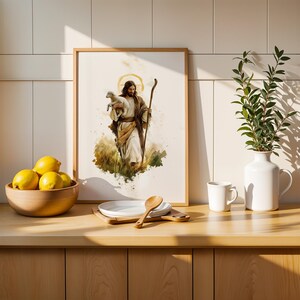 May include: A framed watercolour-style print depicting a figure with long hair and a beard holding a small lamb while carrying a wooden staff. The artwork is displayed on a counter next to a wooden bowl of yellow lemons, a white plate, and a vase.
