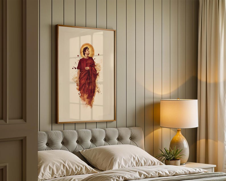 May include: A framed wall art print featuring a figure in a deep red robe with a golden halo, set against a cream background. The artwork displays a watercolour-style aesthetic with scattered dark accents, enclosed in a simple wooden frame.