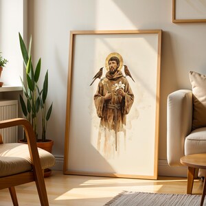 May include: A framed wall art print featuring a watercolor illustration of a bearded figure in a brown hooded robe with a gold halo. The figure holds a white lily, with a small bird perched on each shoulder, set against a light cream background.