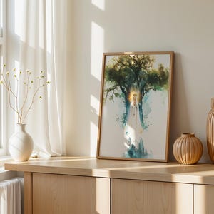 May include: A framed watercolour print featuring a central figure in a white robe standing before a large, dark green tree with a glowing golden halo effect. The artwork is displayed in a light wood frame, positioned on a wooden surface.