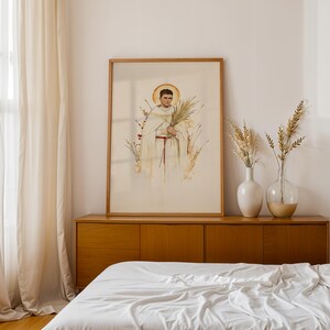 K&ouml;nnte beinhalten: Ein gerahmter vertikaler Wandkunstdruck mit einer Aquarellillustration einer Figur in wei&szlig;em religi&ouml;sem Gewand mit goldenem Heiligenschein, die einen Palmwedel h&auml;lt, umgeben von zartem Trockengras und botanischen Elementen in neutralen T&ouml;nen.
