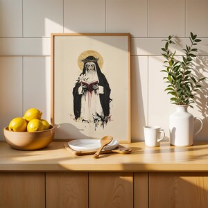 May include: A framed art print featuring a black and white illustration of a figure wearing a crown of thorns and a dark cloak, holding a red book. The print is displayed on a wooden counter next to a bowl of yellow lemons and a white vase with greenery.