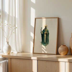 May include: A framed vertical art print featuring a watercolour illustration of a bearded figure in a long green robe and crown, holding a shamrock and a staff, with a golden halo, displayed in a light wood frame on a wooden surface.