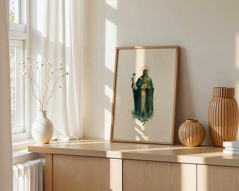 May include: A framed vertical art print featuring a watercolour illustration of a bearded figure in a long green robe and crown, holding a shamrock and a staff, with a golden halo, displayed in a light wood frame on a wooden surface.