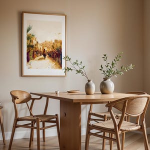 Peut inclure: Une impression d'art encadr&eacute;e repr&eacute;sentant une sc&egrave;ne de foule avec un personnage sur un &acirc;ne, expos&eacute;e au-dessus d'une table &agrave; manger en bois clair avec deux vases en c&eacute;ramique contenant des branches vertes et deux chaises en bois.